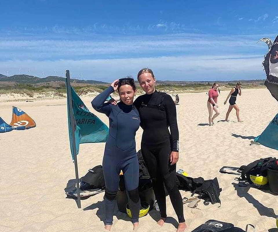 Dos alumnas con trajes de neopreno posan en la playa junto a las banderas de Mysfit Kite School en Tarifa.