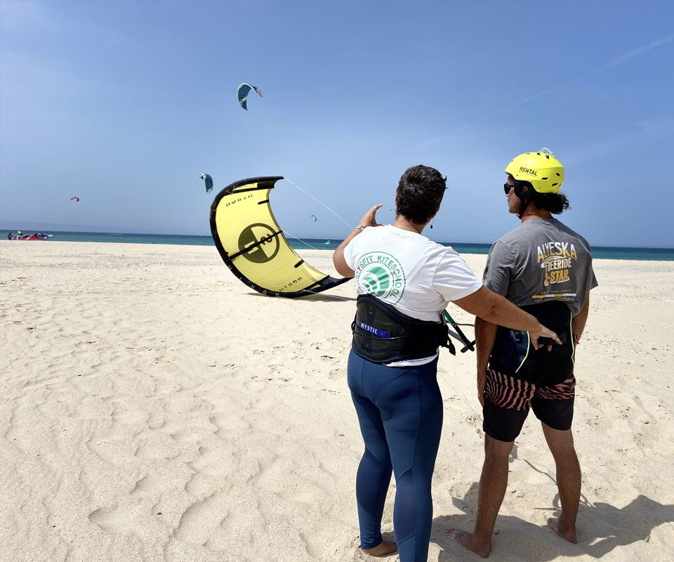 Instructor de kitesurf de la escuela Mysfit Kite School de Tarifa enseñando a un alumno en la playa, con una cometa amarilla parcialmente inflada y varios kitesurfistas navegando en el mar al fondo. Ambos llevan arnés y el alumno un casco amarillo, mientras el instructor da indicaciones señalando el cielo.