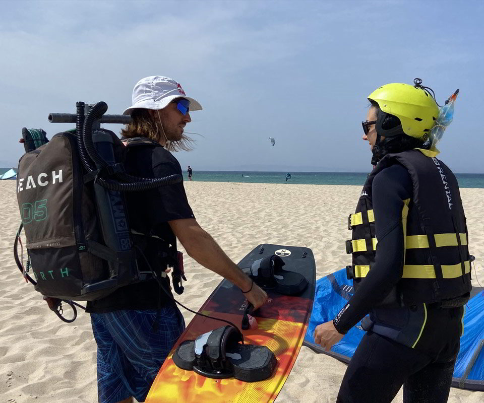 Instructor de kitesurf en la playa de Tarifa explicando a un alumno cómo usar la tabla, ambos equipados con chaleco, casco y material técnico. Al fondo, el mar en calma y varias cometas ya surcando el cielo, junto a una cometa azul en la arena marcada con “REACH 06”.